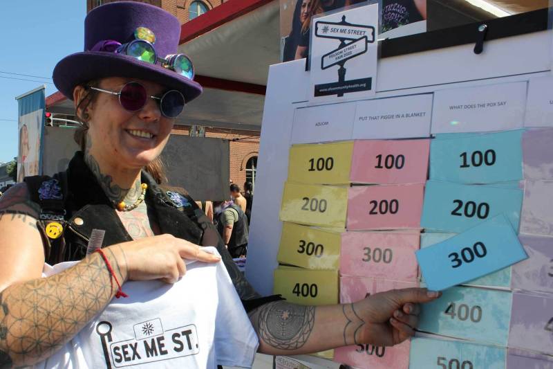 A person wearing a top hat, sun glasses, and a black vest with several pins, holds up a white t-shirt in one hand and points to a board with different colored numbers in the other hand.