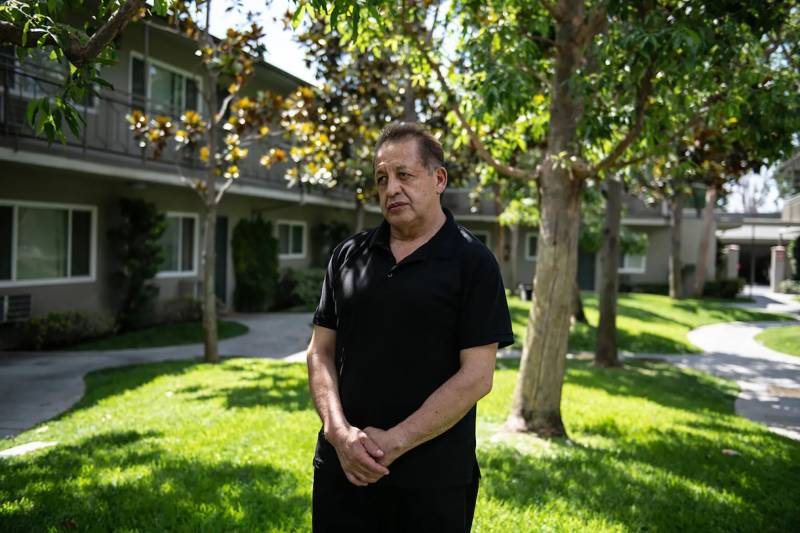 In the center of the frame stands a middle-aged Latino man with short, swept-back, dyed brown hair, his hands clasped in front of him, wearing a black short-sleeved polo shirt and black trousers. He looks a bit forlornly to the right of the camera. Beyond him is a grassy lawn in the center of a two-story apartment complex, with dappled sunlight lighting the green through the leaves of multiple deciduous trees.