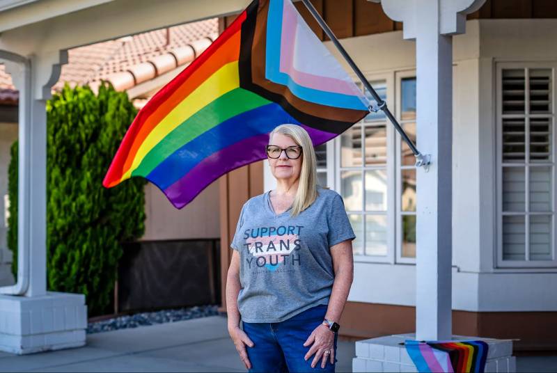 woman with straight blonde hair and glasses wearing 'support trans youth' shirt stands underneath a pride flag outdoors outside of a home