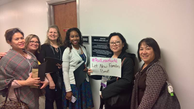 Six women of different races smile at the camera and one of the women holds up a hand-written sign that reads "#Paid Leave For All Let New Families Bond"