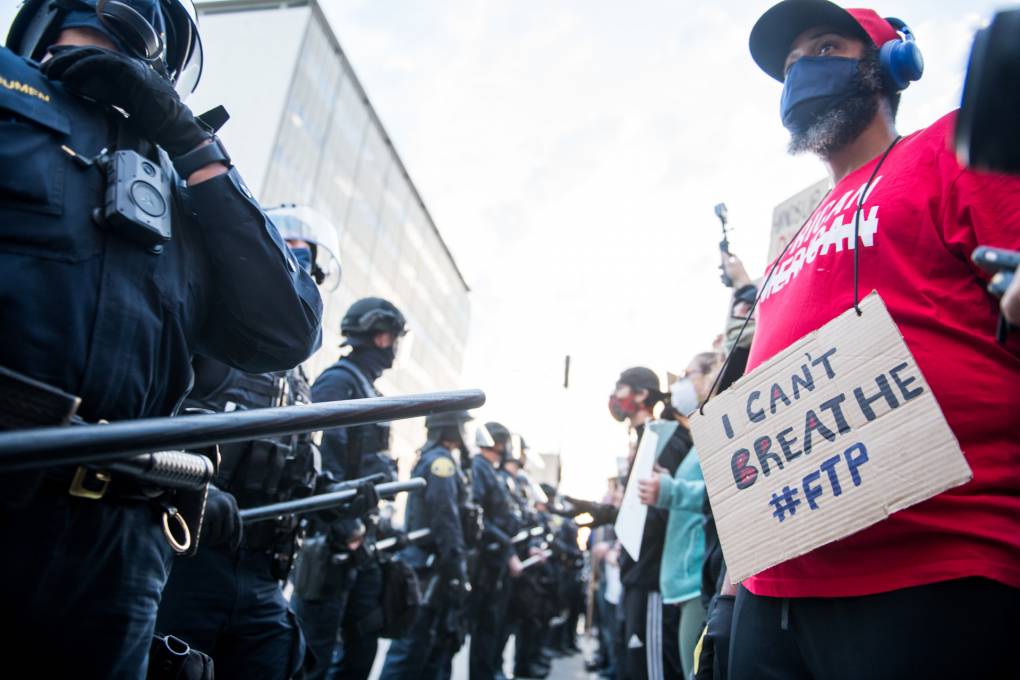 a row of police officers in combat gear faces a row of protestors, one holding a sign that says 'I can't breathe #FTP'