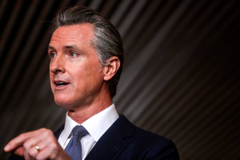 Governor Gavin Newsom is wearing a navy suit and blue tie, looking away from the camera with finger pointing as he speaks.