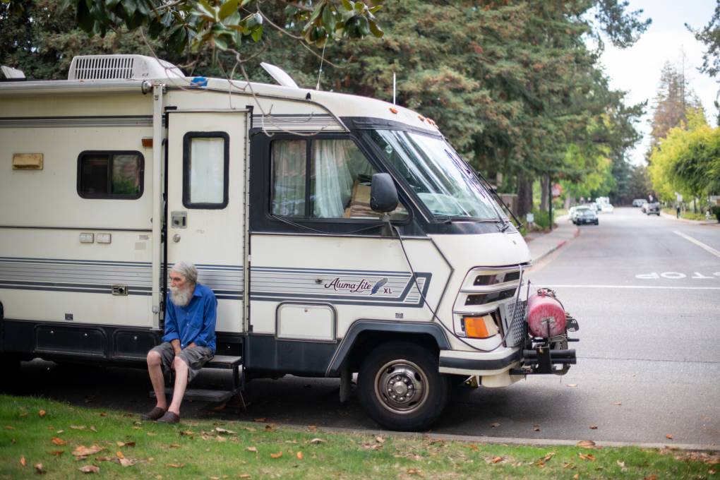 elderly man in blue shirt with large white beard sits on step outside an RV truck parked at the side of a street