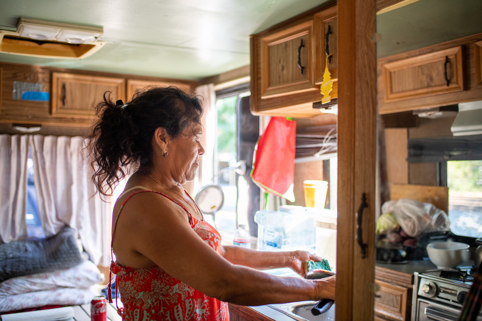 woman wearing red dress washes dishes at a sink inside an RV as light streams in through the window