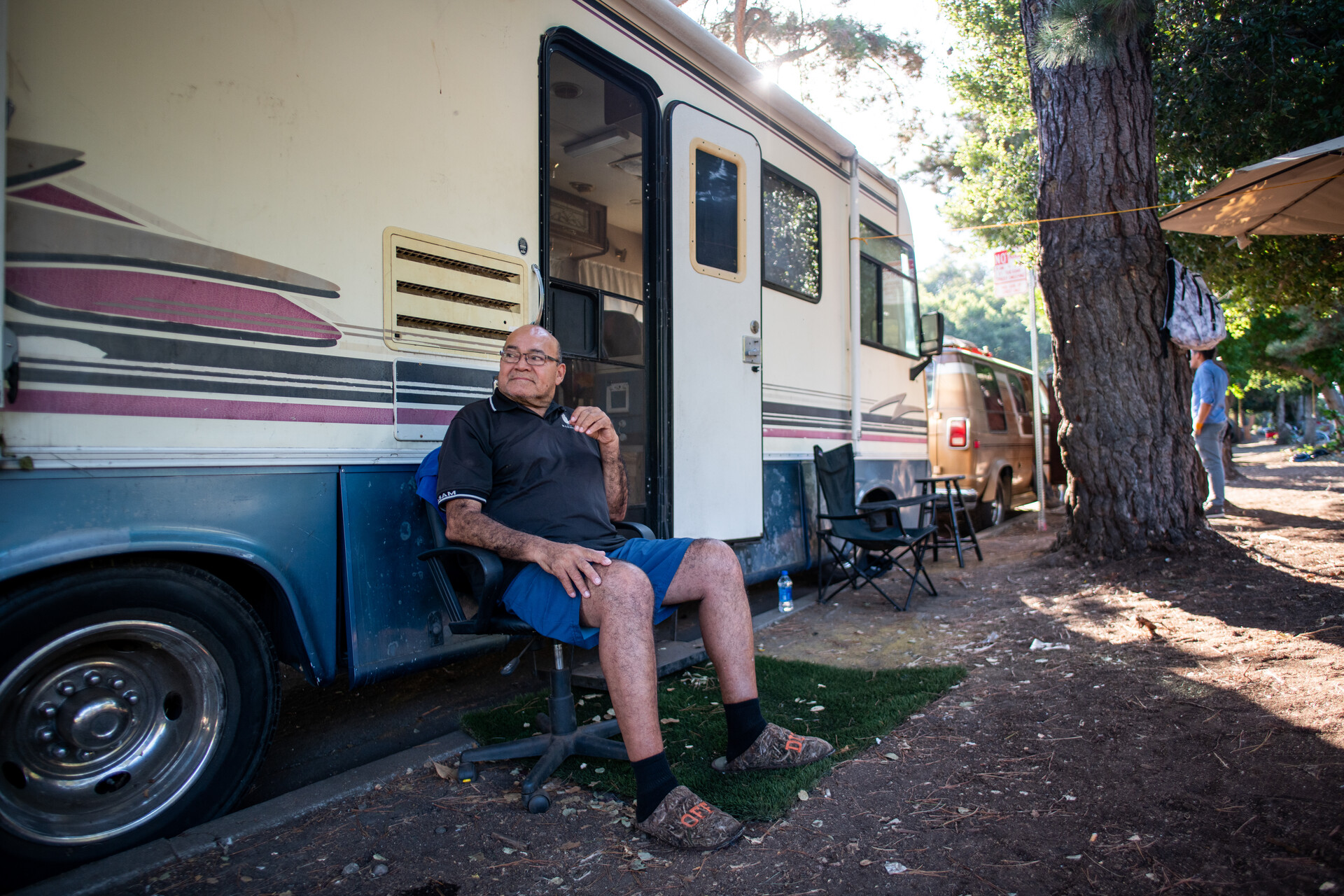 smiling man wearing blue shorts sits in a folding chair outside the side door of his RV