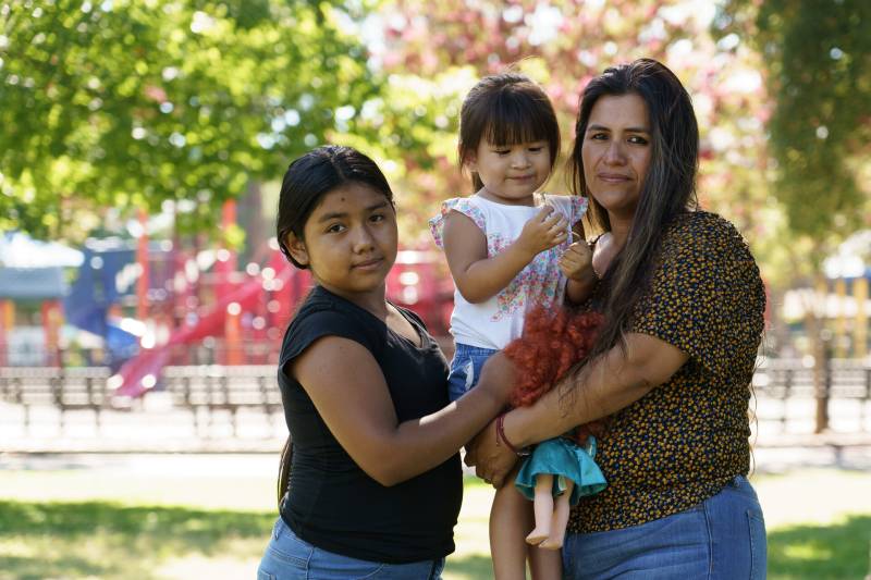 A Latina woman holding a 2-year-old daughter next to her 9-year-old daughter looking at the camera in a park with a playground in the background on a sunny day