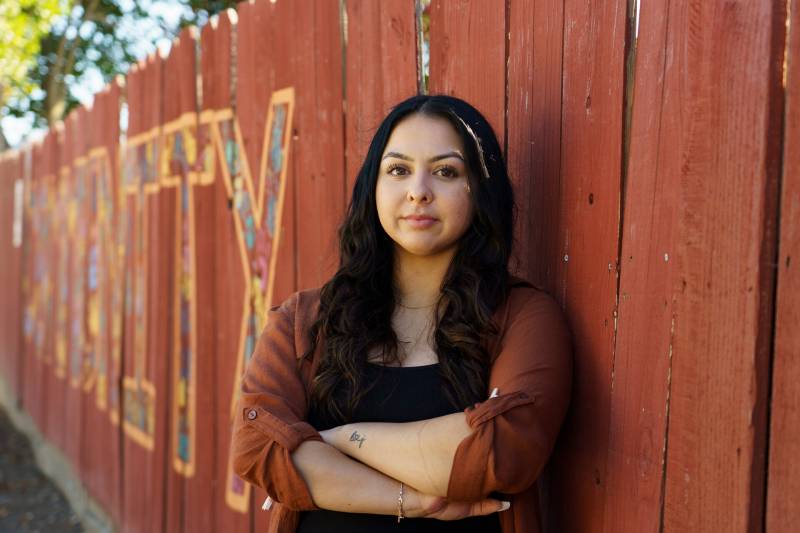 A young Latina woman of 20 with long black hair stands with her arms folded in front of a red wooden fence as she looks at the camera with a determined expression.