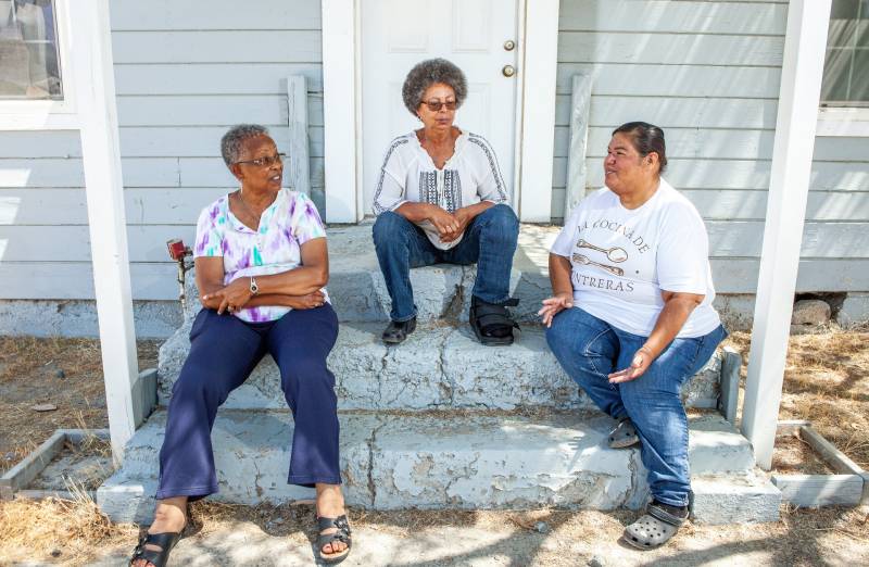 Two Black women with natural salt-and-pepper hair and one Latina woman with black hair pulled into a tight bun sit on the cracked cement front steps of a gray, wood-paneled building with a white door. The women wear blue jeans. One woman wears a white t-shirt with front buttons and splashes of purple and green. One woman wears a white cotton shirt with black embroidery down the front and arms. The third woman wears a white t-shirt reading "La Cocina de Contreras."