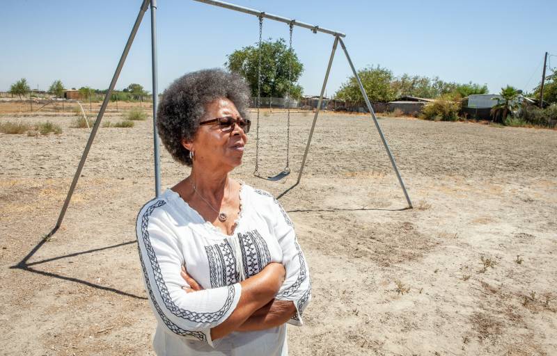 A woman with coffe-and-cream skin, sunglasses and gray-speckled natural black hair stands with arms folded in front of a swingset tha thas one swing. She wears a white cotton top with a v-neck and black geometric embroidery down the arms and front. The ground is bare, dry soil. A few trees are in the distant background.