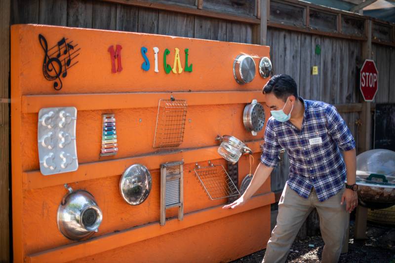A man in a blue plaid shirt and surgical mask, with grey pants stands in a yard and gestures toward a bright orange wall adorned with varies items nailed or fixed in place such as pots, pans, a colander and a xylophone. Above them all on the wall is the word musical. 
