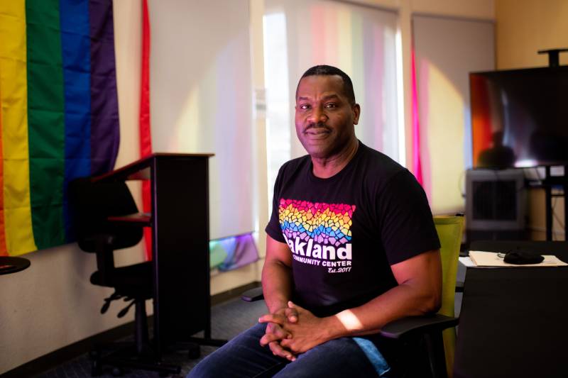 A man wearing a black shirt with rainbow colors in the middle spelling "Oakland" sits down inside a building.