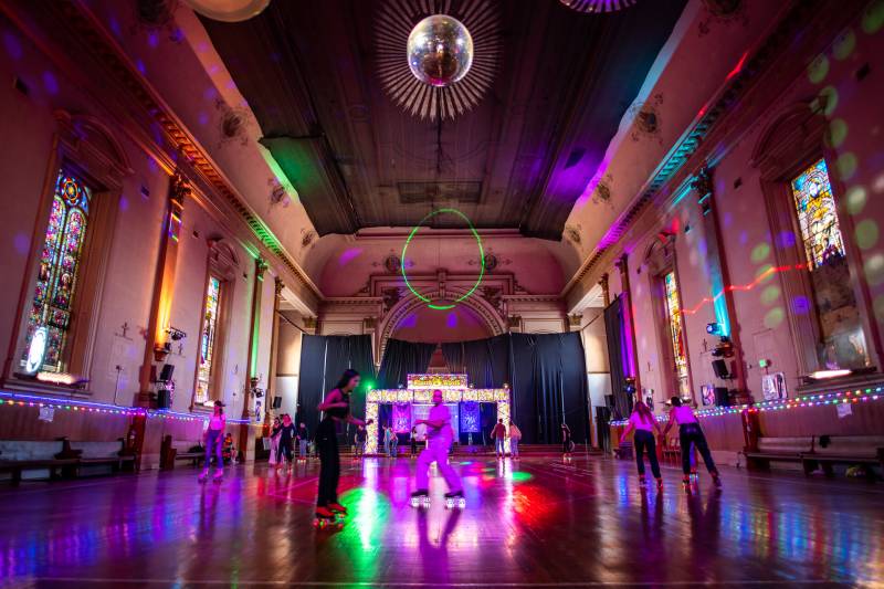 People in roller skates cruise around a large empty church, now converted into a roller rink. Multicolored laser lights fill the room, and a shimmering disco ball hangs from the ceiling. The walls of the room are lined with the church's original stained glass windows depicting Catholic religious imagery.