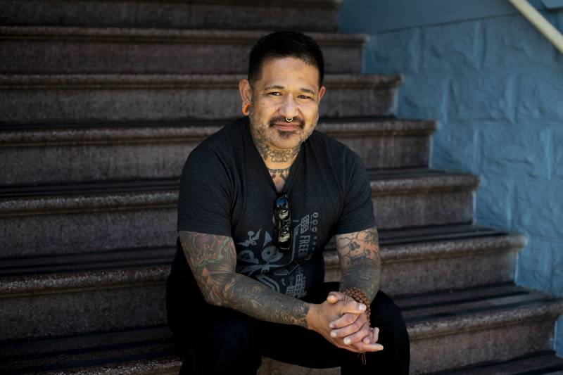 A man wearing a short sleeved black shirt with tattoos on his arms and a nose ring sits on steps with his hands together.