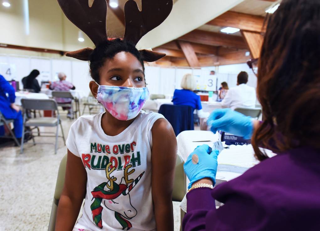 A healthcare worker crouches in front of the camera out of focus, preparing to give a vaccine to a Black child wearing a holiday themed shirt and reindeer antlers