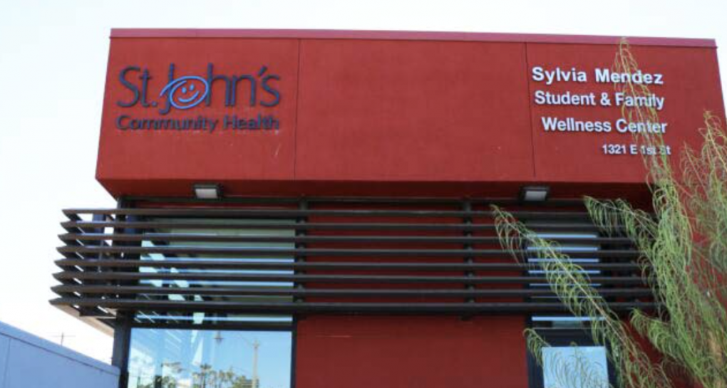 The image shows a brick-red building with tall windows. On the left of the building, black lettering reads "St. John's Community Health" with the "O" as a smiley face. On the right side of the building, white lettering reads, "Sylvia Mendez Student and Family Wellness Center."