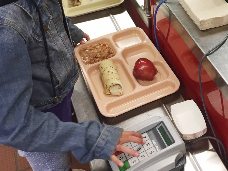The image shows the hand of a student with white skin, wearing a jean jacket, punching a number into an electronic payment system. On a tray at the center of the image is an apple, burrito, and baked beans.