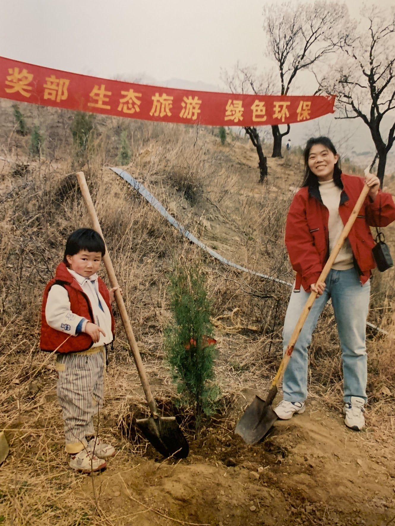 a young child and and their mother plant a tree on a hillside with a banner with Chinese characters hanging behind them