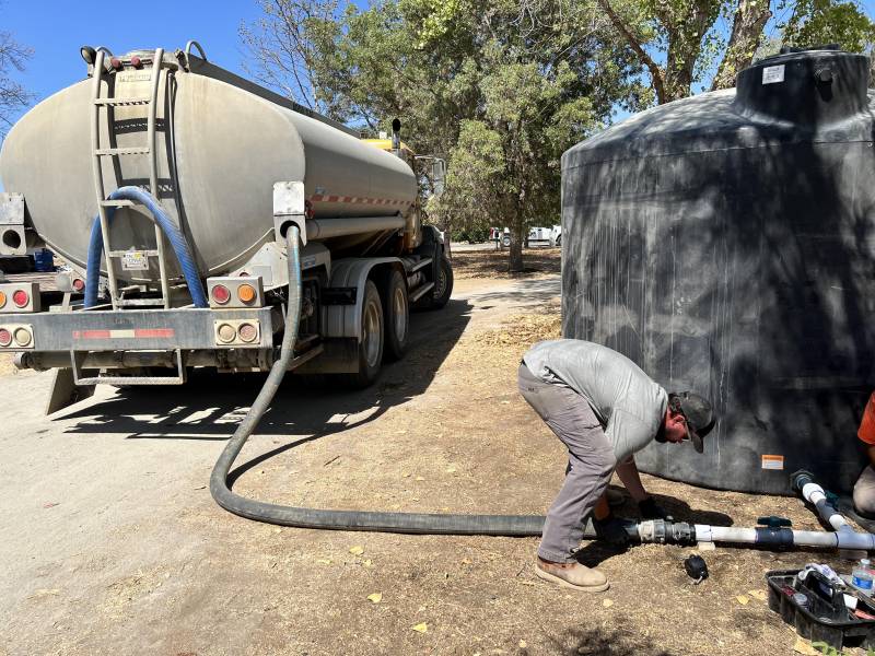 A man bends over in front of a water tank outside somebody's home as he tries to attach a large hose connected to a water tanker seen from the rear in the background with a dirt road leading away to a dry landscape and a tree in the background.