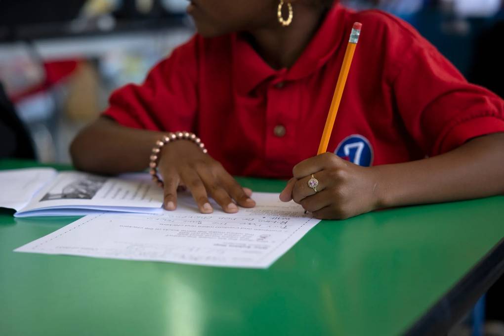 A young Black child wearing a red shirt, sitting at a green desk taking a test with a pencil in hand