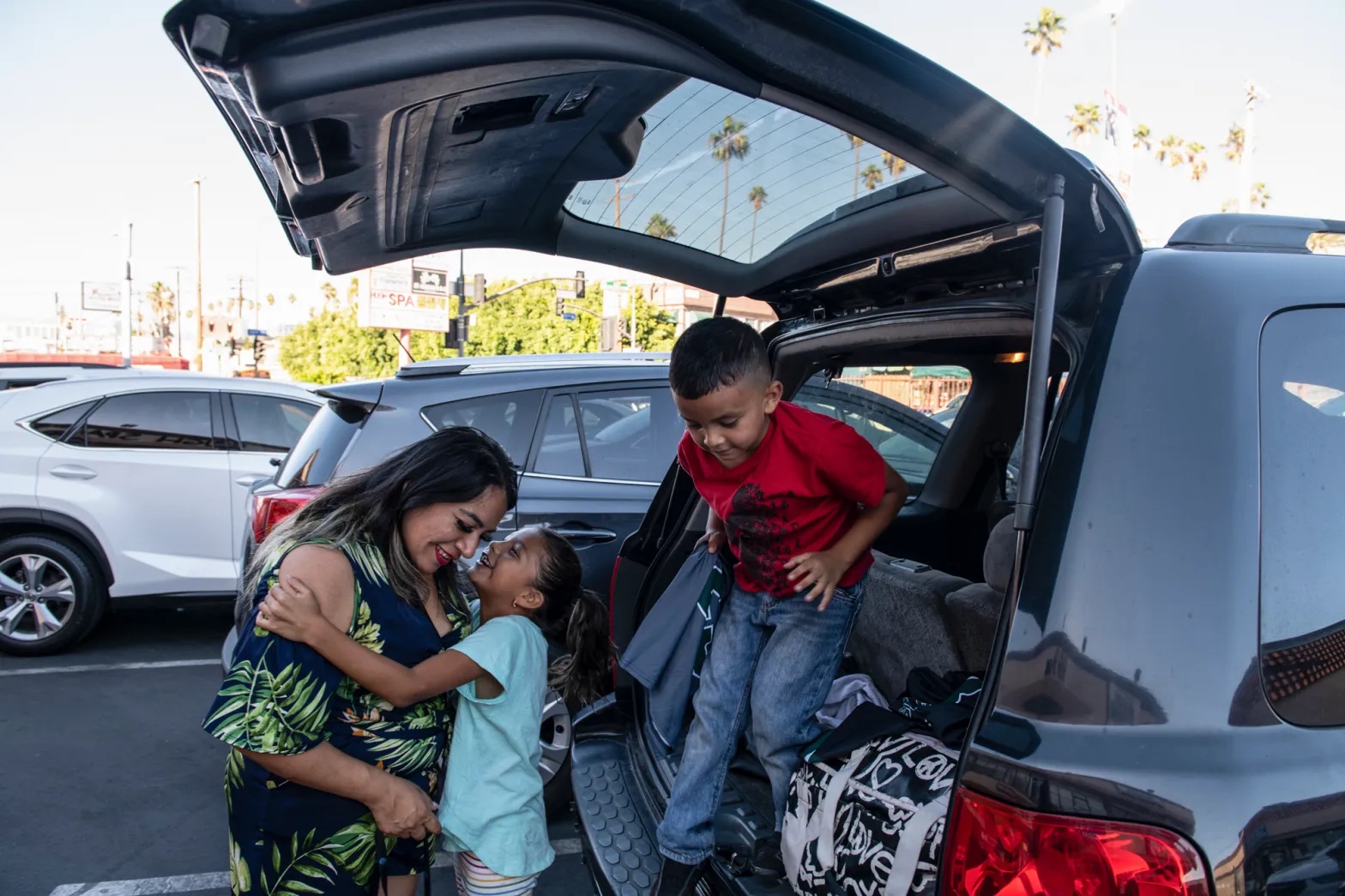 A Latina woman helps her two small kids out of the car, one of the kids is kissing her on the cheek as the other gets out.