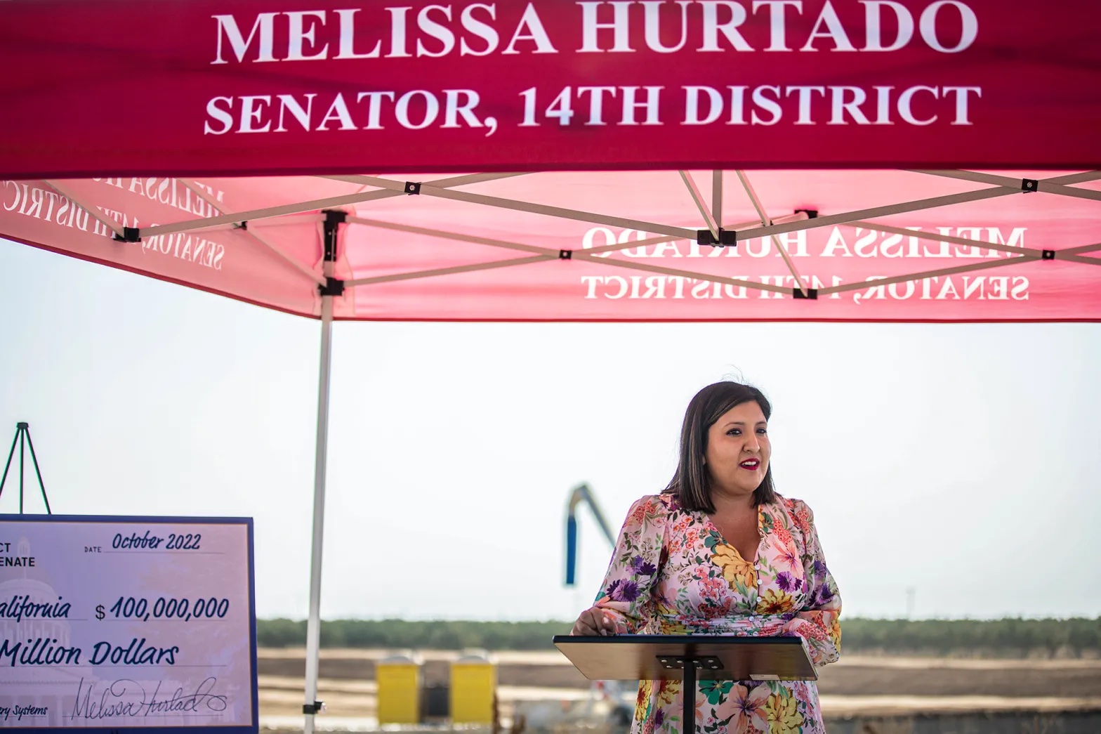 A Latina woman speaks under a fold-up tent that has "Melissa Hurtado, Senator, 14th District" written on it.