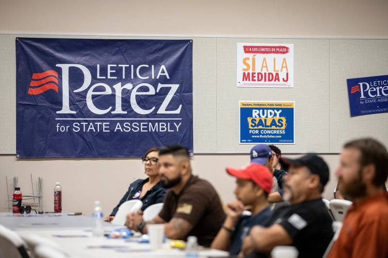 Four people sit behind a table with a poster on the wall behind that reads "Leticia Perez - State Assembly"