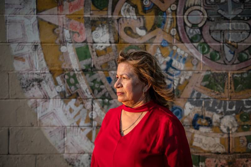 an older Latina woman in a red shirt stands in front of a mural