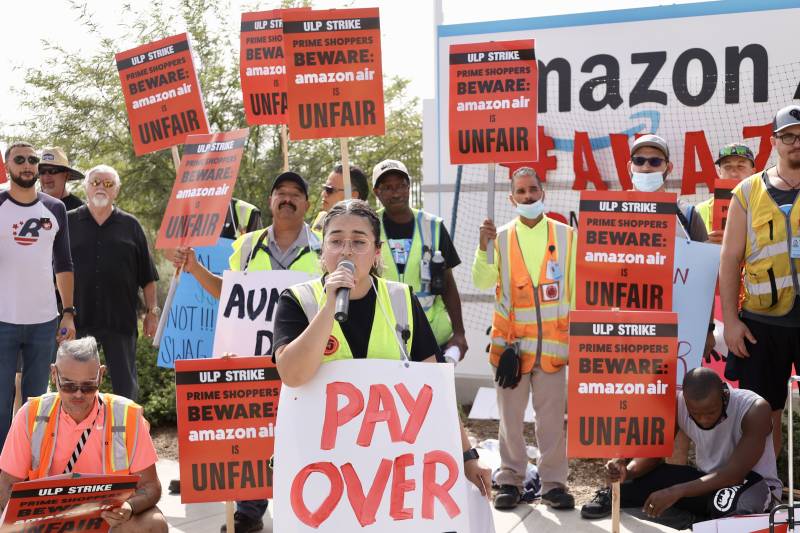 Striking workers carry signs as a woman speaks into a microphone holding a sign that says "pay over"