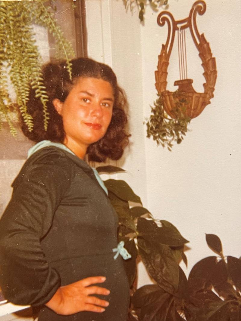 An aged photograph of a young woman with dark wavy hair in a dark green shirt. She stands sideways with a hand on one hip, looking at the camera. There are plants around her and a lyre on the wall.