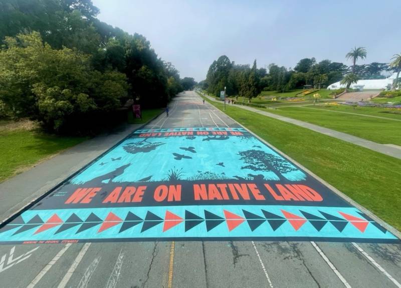A massive turquoise, black and red street mural covering a section of the JFK Promenade stating 'We are on Native land'