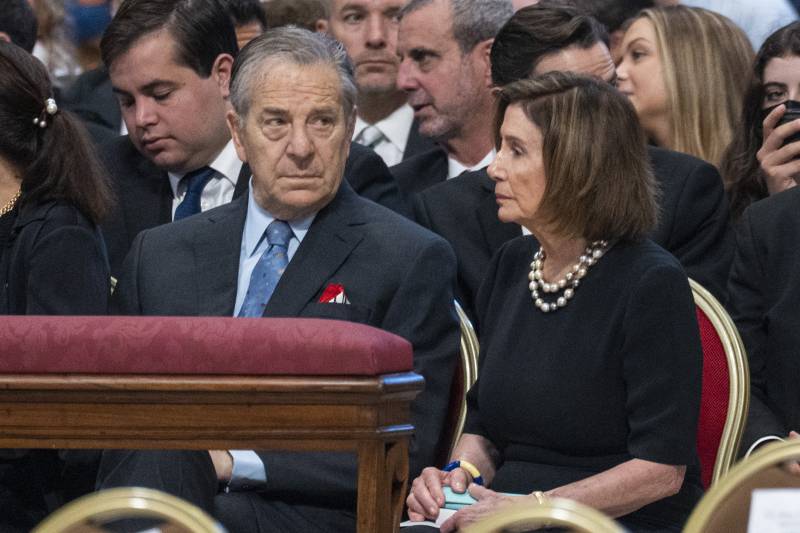 Nancy and Paul Pelosi sit in chairs, both wearing dark colored formal attire.