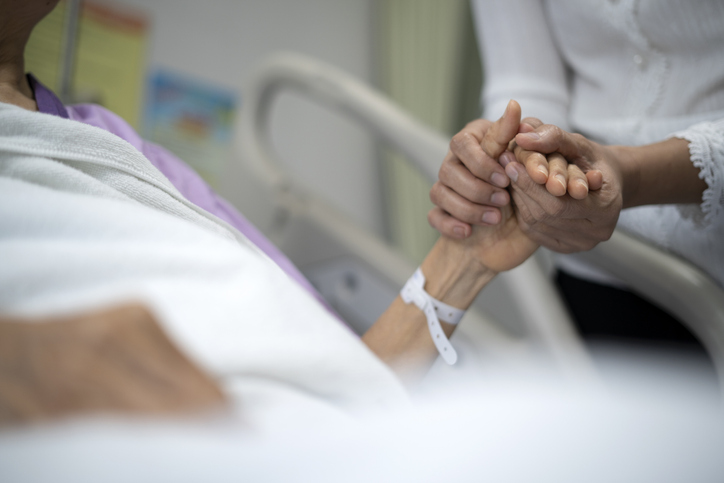 A woman lying in a hospital bed holds hands with a loved one.