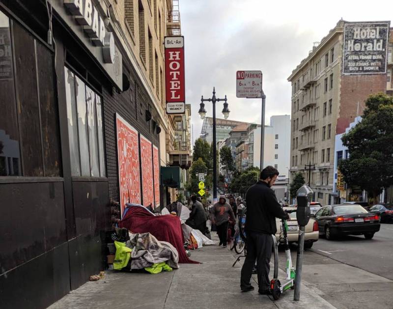 A man stands in the foreground of a street in San Francisco's Tenderloin district with homeless encampments lining the sidewalk.