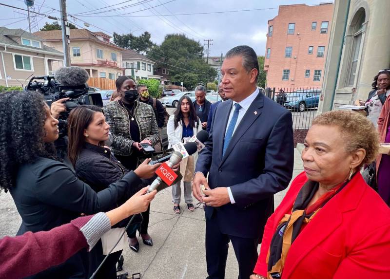 An older man in a blue suit and tie and an older woman in a red jacket talk to reporters on the street outside of a church.