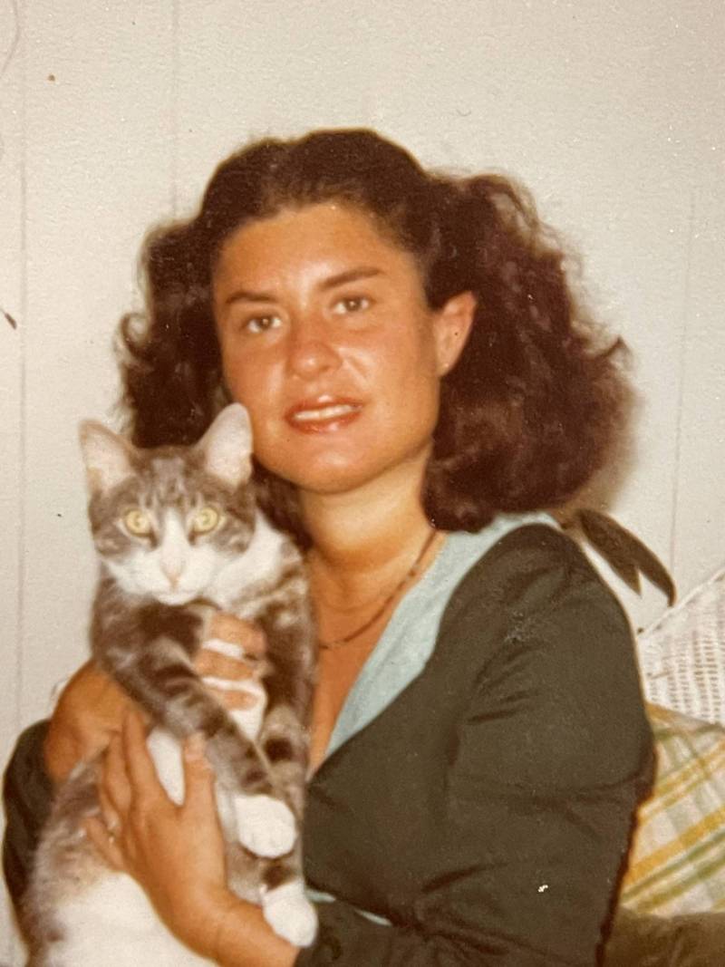 An aged color photograph of a woman with wavy brown hair. She is holding a grey and white striped cat. They are both looking at the camera. 