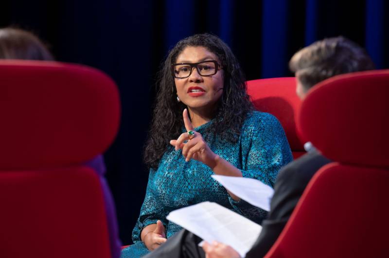 Mayor London Breed sits in a red chair on a stage, wearing black framed glasses, holding her hand up with her pointer finger up as if to articulate a point. The backs of two chairs can be seen at either side of her, out of focus, in the foreground.