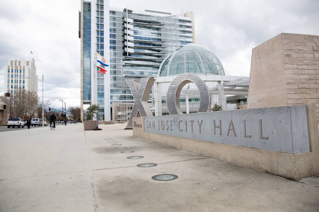 A view of San Jose City Hall, a large modern white 15-story building with a glass dome in the foreground and a sign out front that says "San Jose City Hall"