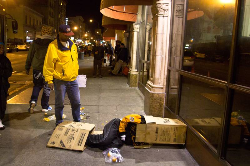 A worker in a yellow jacket looks on at an unhoused person sleeping in a cardboard box on the street