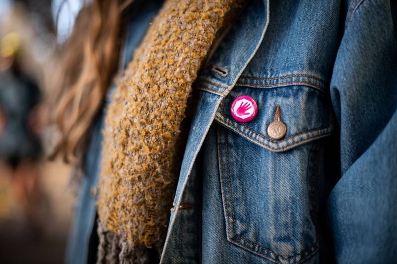 The jacket of a girl with part of a scarf draped over the front and a pink round pin with a white background and a pink hand splayed out.
