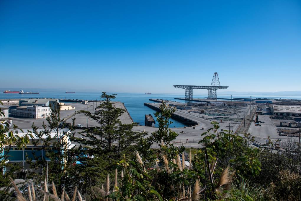 A view from a hill of a dilapidated shuttered and abandoned naval shipyard with a dock and a rusting equipment, bushes in the foreground, the blue sea and sky past the dock looking out at San Francisco Bay