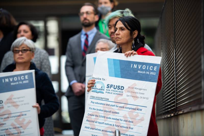 A woman holds a sign that looks like an invoice written out to SFUSD