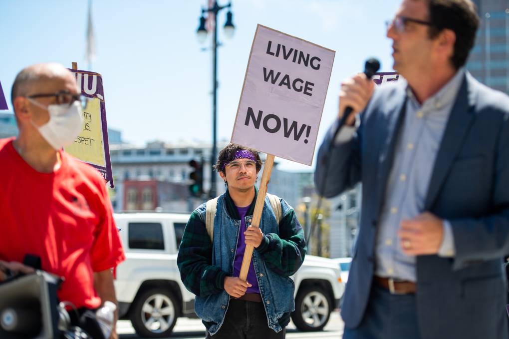 three people at a rally, with one holding a sign that reads 'living wage now!' and one speaking into a microphone