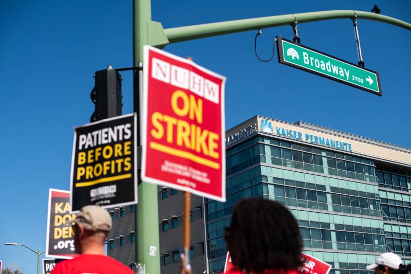 Workers on the street hold signs that say 'Patients Before Profits' and 'On Strike,' with the Kaiser Permanente building in the background.