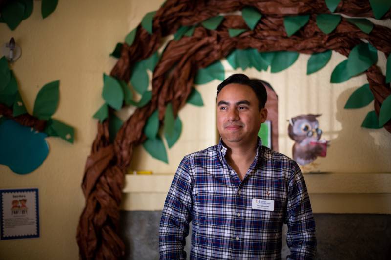 a Latino man in a blue and white checked shirt poses for a portrait in front of a colorful tree decoration in an office