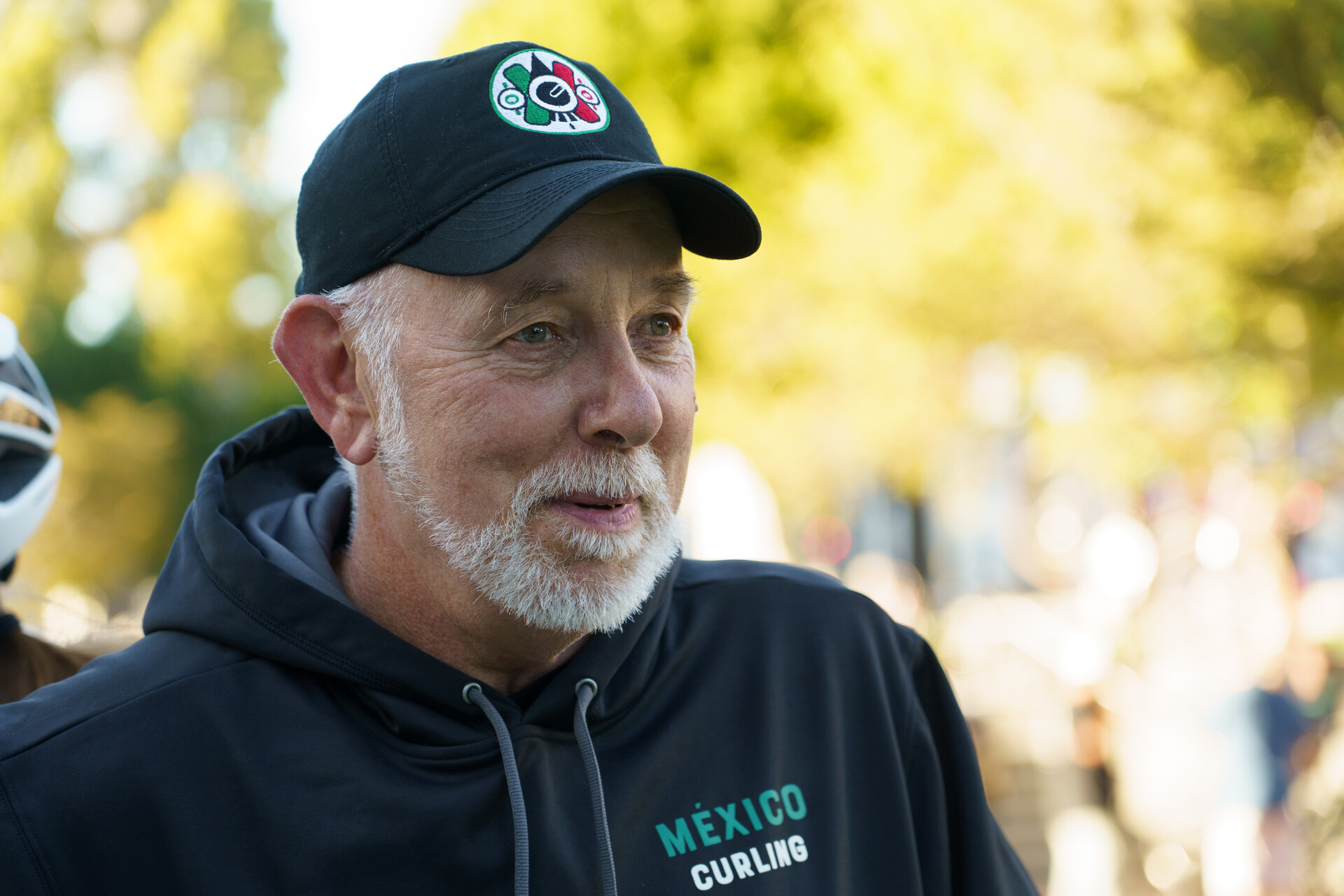 A man in his 60s with a trimmed white beard wearing a black cap and a black hoodie smiles as he looks away from the camera.
