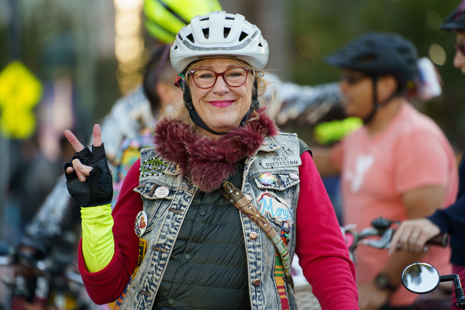 A woman in her seventies wearing a helmet, glasses, red long-sleeve sweater and a jean vest waves at the camera.