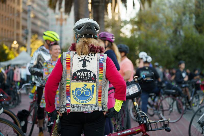 A woman in her 70s with her back turned to the camera reveals political messages in support of cycling and water conservation with other cyclists in the background.