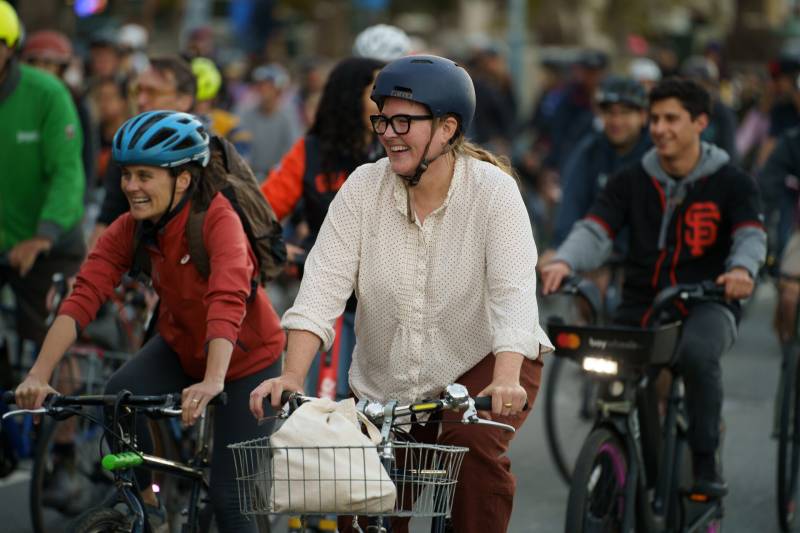 A woman in a white shirt and a woman in a red shirt with helmets and riding bikes, smile with other cyclists around them.
