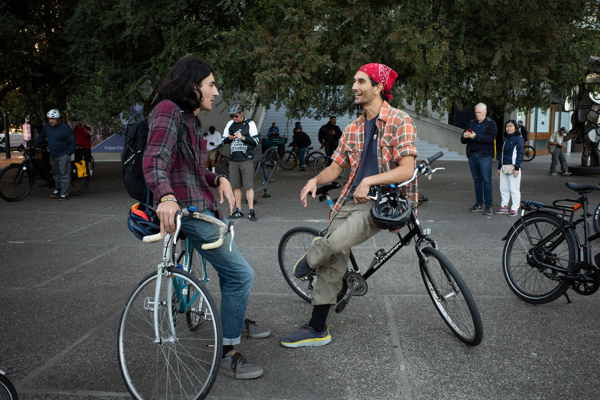 Two men leaning on their bikes have a chat, one has long hair, the other is wearing a red bandana, both are in the early 20s.