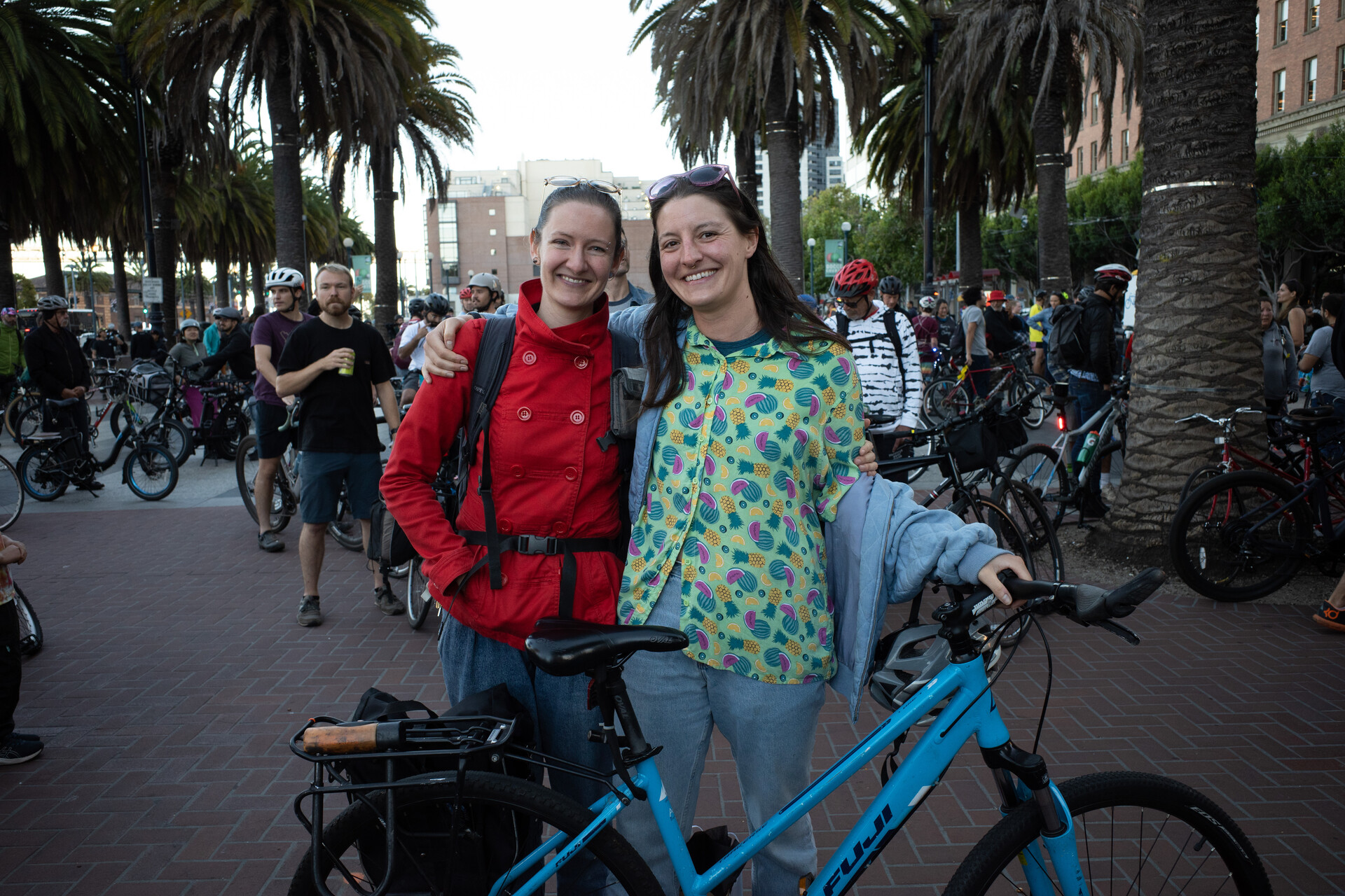 Two women in their 30s pose for the camera in front of a blue bike, one woman is wearing a red jacket, the other a green one with patterns, both are smiling.